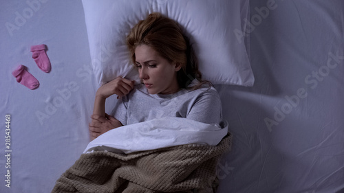 Unhappy woman lying in bed with baby socks beside  baby expectation  infertility