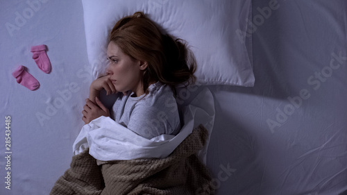Sad lady crying lying in bed and looking at baby socks near pillow, miscarriage