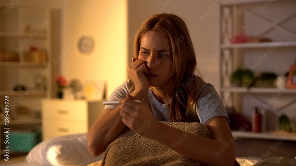 Worried female holding pregnancy test sitting on bed, infertility problems
