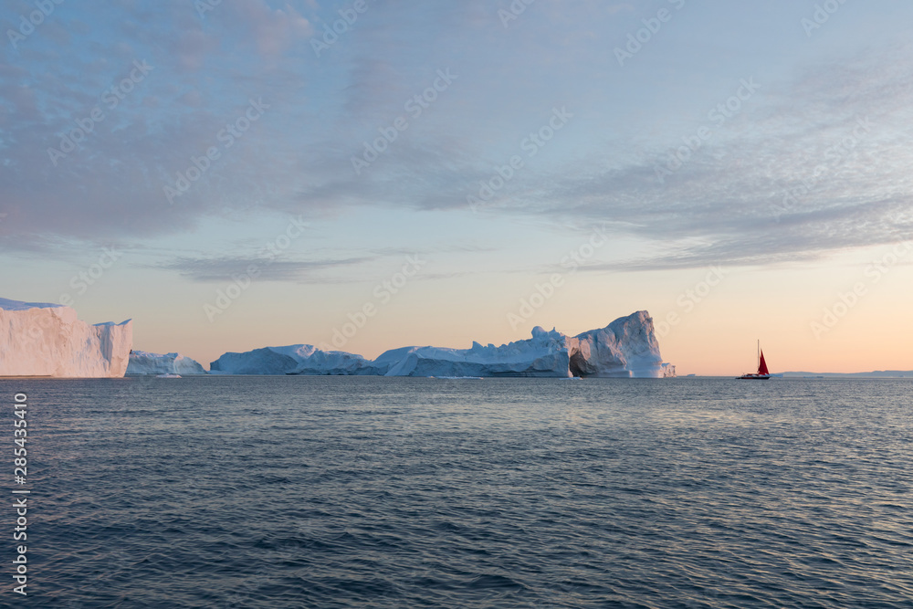A small boat among icebergs. Sailboat cruising among floating icebergs in Disko Bay glacier during midnight sun Ilulissat, Greenland. Studying of a phenomenon of global warming Ices and icebergs