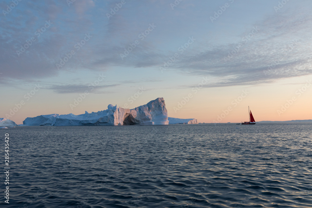 A small boat among icebergs. Sailboat cruising among floating icebergs in Disko Bay glacier during midnight sun Ilulissat, Greenland. Studying of a phenomenon of global warming Ices and icebergs A small boat among icebergs. Sailboat cruising among floating icebergs in Disko Bay glacier during midnight sun Ilulissat, Greenland. Studying of a phenomenon of global warming Ices and icebergs
