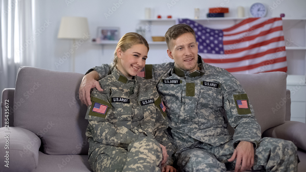 Cheerful couple of soldiers sitting on sofa against US flag, happy homecoming