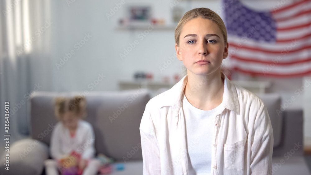 Fototapeta premium Sad single mother looking at camera daughter playing on sofa against flag of USA
