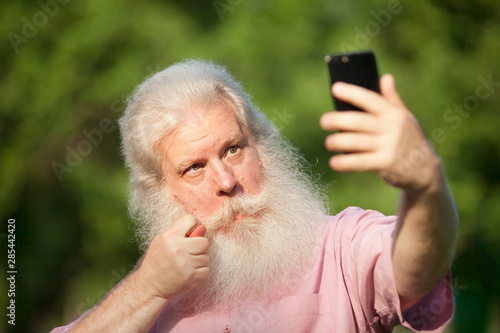 Bearded senior man in casual wear making selfie on smartphone and showing fig sign, standing against blur green natural background in city park at sunny day. photo