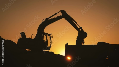 Silhouette of an excavator that loads sand into a truck at sunset. Concept construction and heavy industry, machine will be used in heavy industry business. Slow motion footage.