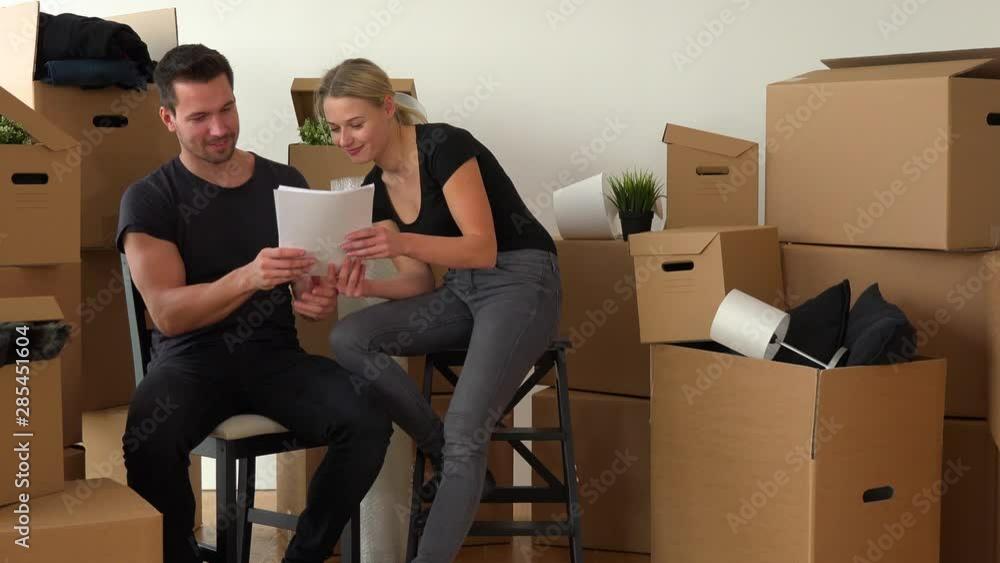 A happy moving couple sits on chairs in an empty apartment and reads a bunch of papers, surrounded by cardboard boxes A happy moving couple sits on chairs in an empty apartment and reads a bunch of papers, surrounded by cardboard boxes