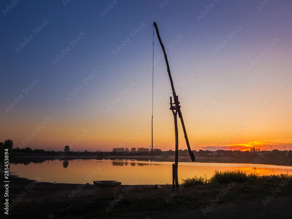 Idyllic rural scene with an old water well (popular called the stork well in the old villages of Moldova, also well sweep or shadoof). Calm sunset sky reflecting on the lake water surface.