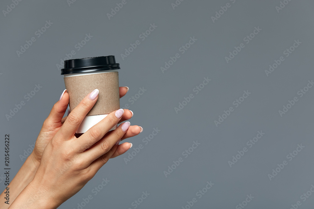 A beautiful female hand offers a hot drink, tea or coffee in a disposable natural glass on a light gray background. CONCEPT start of the day with a cup of coffee