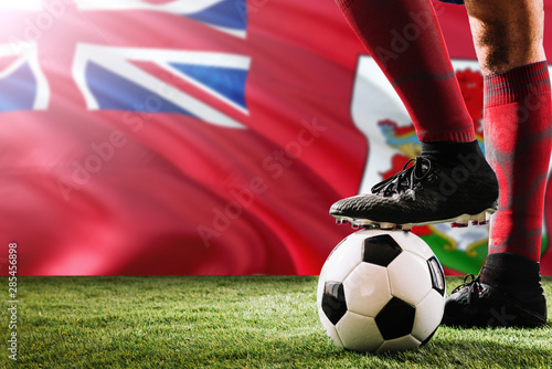 Close up legs of Bermuda football team player in red socks, shoes on soccer ball at the free kick or penalty spot playing on grass.