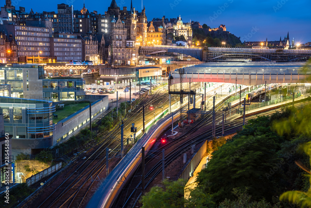 View from above of a train leaving Edinburgh train station at night. Motion blurred. Edinburgh old town skyline is in background.
