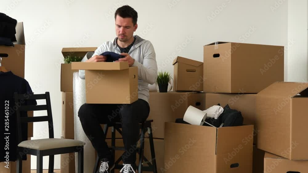 A moving man sits on a chair and puts things into a cardboard box in an empty apartment, surrounded by other boxes A moving man sits on a chair and puts things into a cardboard box in an empty apartment, surrounded by other boxes