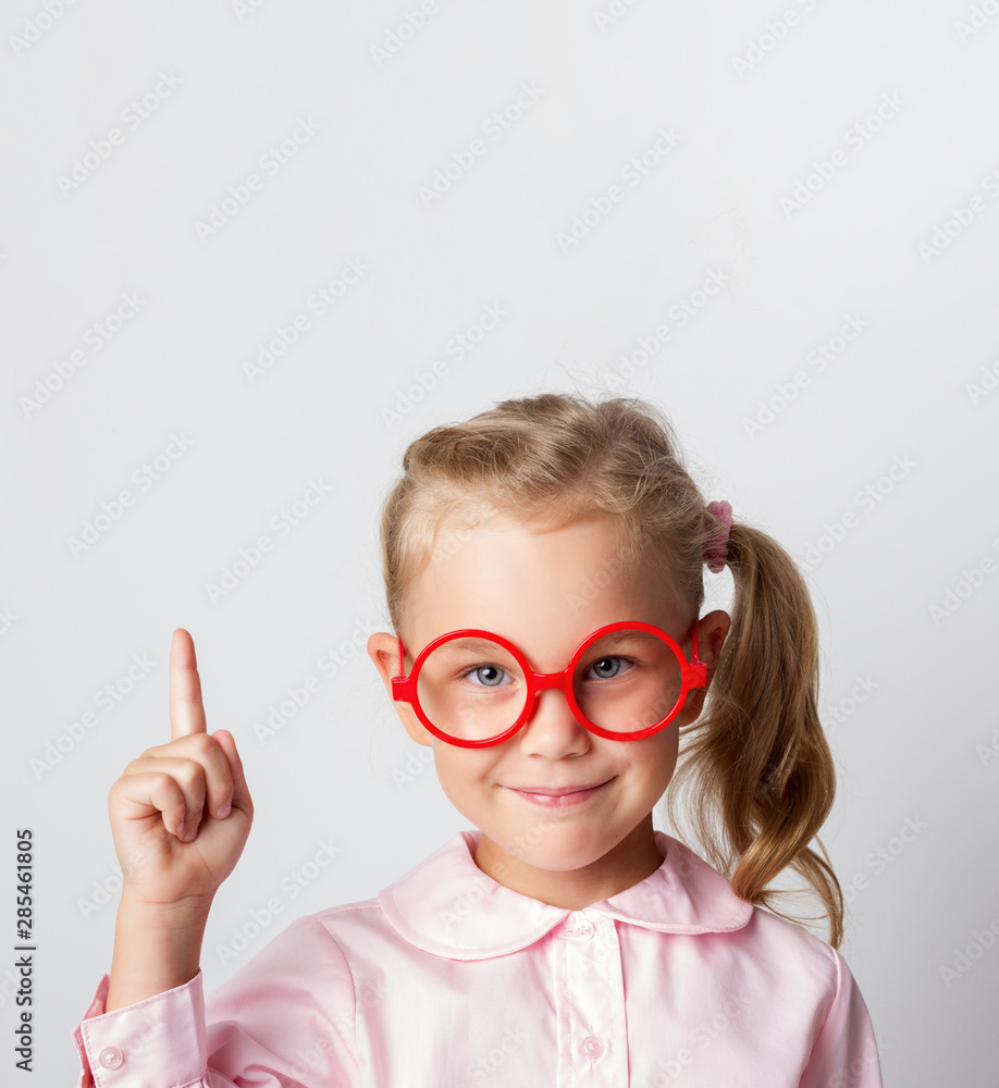 Close-up portrait of amazed pretty young girl schoolgirl in red glasses and a light blouse, pointing finger up, looking at camera with a smile, isolated on a light background