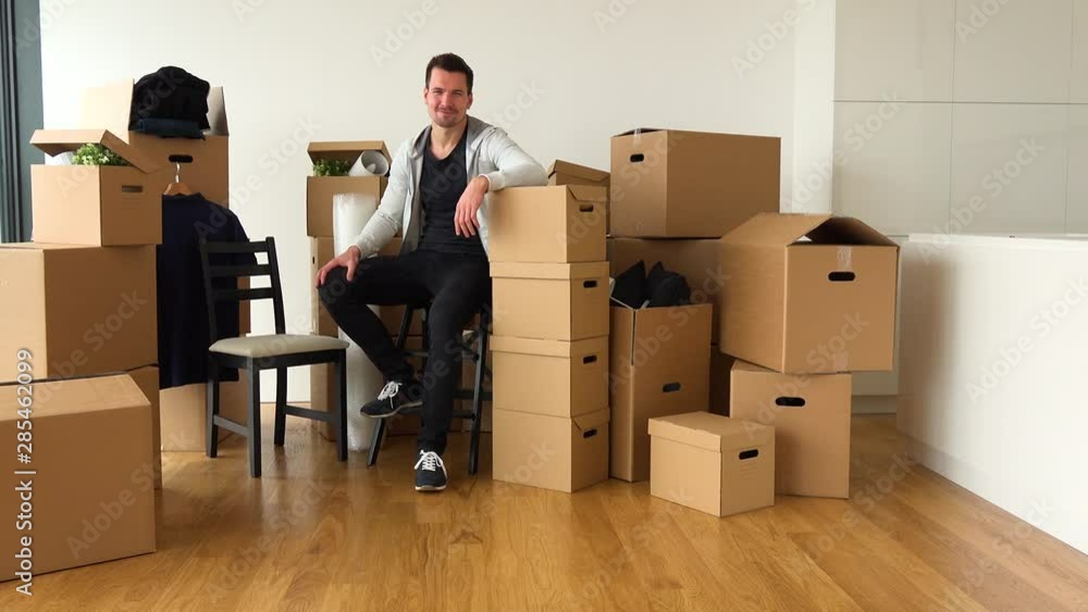 A happy moving man sits on a chair and smiles at the camera in an empty apartment, surrounded by cardboard boxes A happy moving man sits on a chair and smiles at the camera in an empty apartment, surrounded by cardboard boxes