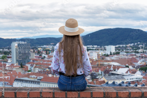 A slender girl with long hair in a hat sits on the edge of a tall building and looks at the city of Graz.