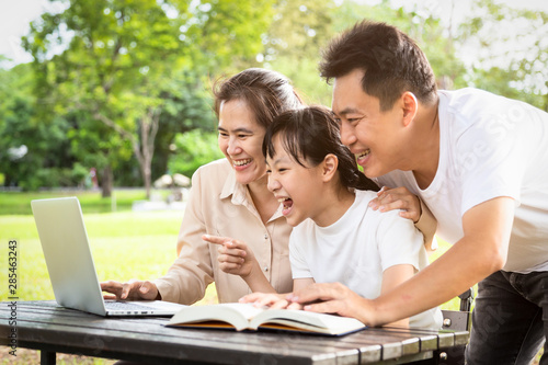 Happy asian family,father,mother,daughter enjoying,smiling using laptop computer in park,parents and child girl having fun playing game or watching video,movie,social networks,media addiction concept