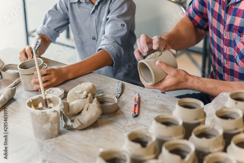 Close up shot of middle aged caucasian man and little boy work together with clay cups in pottery workshop.