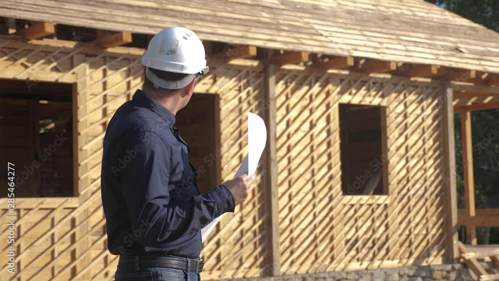 concept building constructing architect slow motion video. man builder in a helmet stands at a construction holding a scheme house plan. site near a wooden frame house under lifestyle construction concept building constructing architect slow motion video. man builder in a helmet stands at a construction holding a scheme house plan. site near a wooden frame house under lifestyle construction