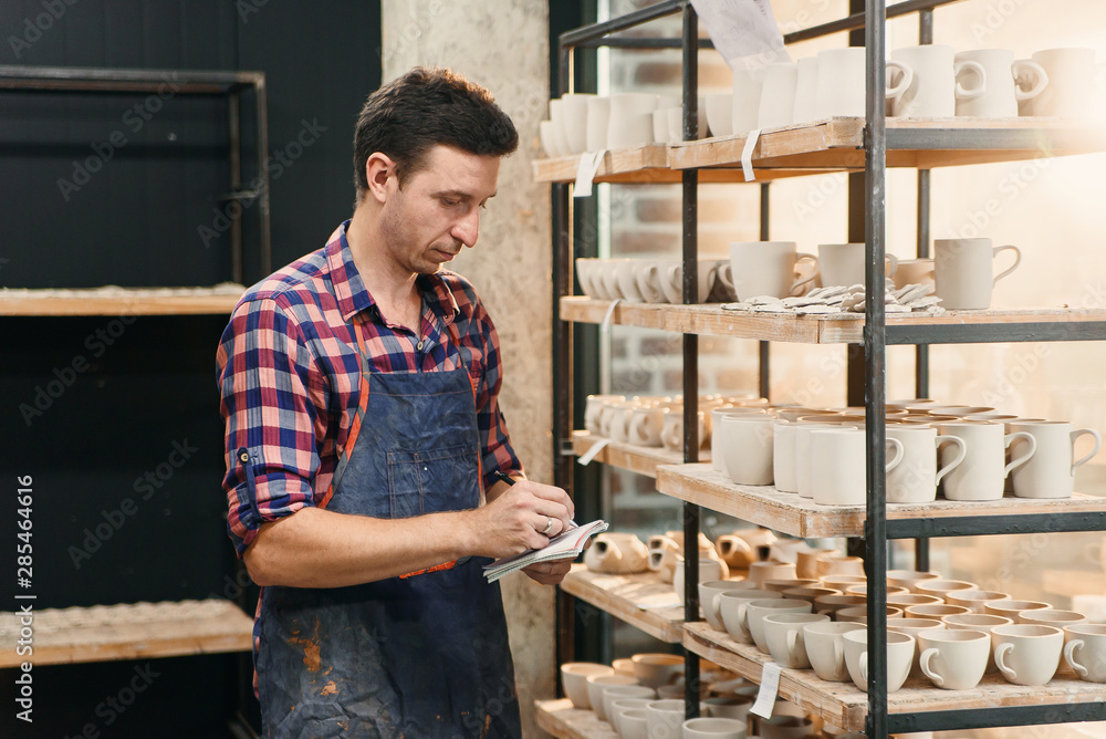 Middle aged male potter in apron notes something in his notebook while counts pottery on shelves. Middle aged male potter in apron notes something in his notebook while counts pottery on shelves.