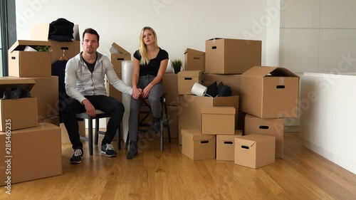 A moving couple sits on chairs, holds hands and looks seriously at the camera in an empty apartment, surrounded by cardboard boxes