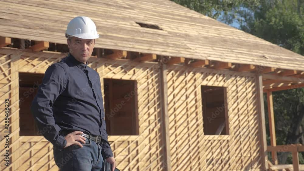 concept building constructing architect slow motion video. man builder in a helmet stands at construction holding digital tablet a scheme house plan. site near a wooden frame house under construction concept building constructing architect slow motion video. man builder in a helmet stands at construction holding digital tablet a scheme house plan. site near a wooden frame house under construction