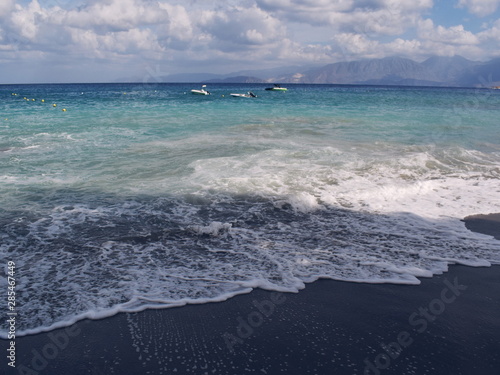 A foamy wave runs onto a sandy beach, the sun through the clouds illuminates the sea, Crete, Greece