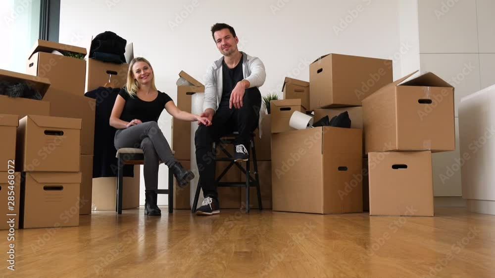 A happy moving couple sits on chairs in an empty apartment, holds hands and smiles at the camera, surrounded by cardboard boxes A happy moving couple sits on chairs in an empty apartment, holds hands and smiles at the camera, surrounded by cardboard boxes