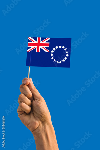 Woman hand holding Cook Islands flag with stick, waving flag on deep blue sky. National theme, deep blue sky. © sezerozger
