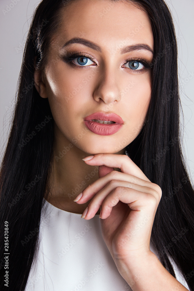 Close-up portrait of sensuality young brunette girl with blue eyes, woman with stylish make up is looking straight and smiling cute with hand near her face on gray wall background, beauty concept Close-up portrait of sensuality young brunette girl with blue eyes, woman with stylish make up is looking straight and smiling cute with hand near her face on gray wall background, beauty concept