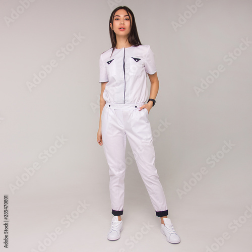 young stylish happy nurse with long brunette hair is standing with hand in her pocket, looking straight and smiling on white wall background. medical fashion concept. free space