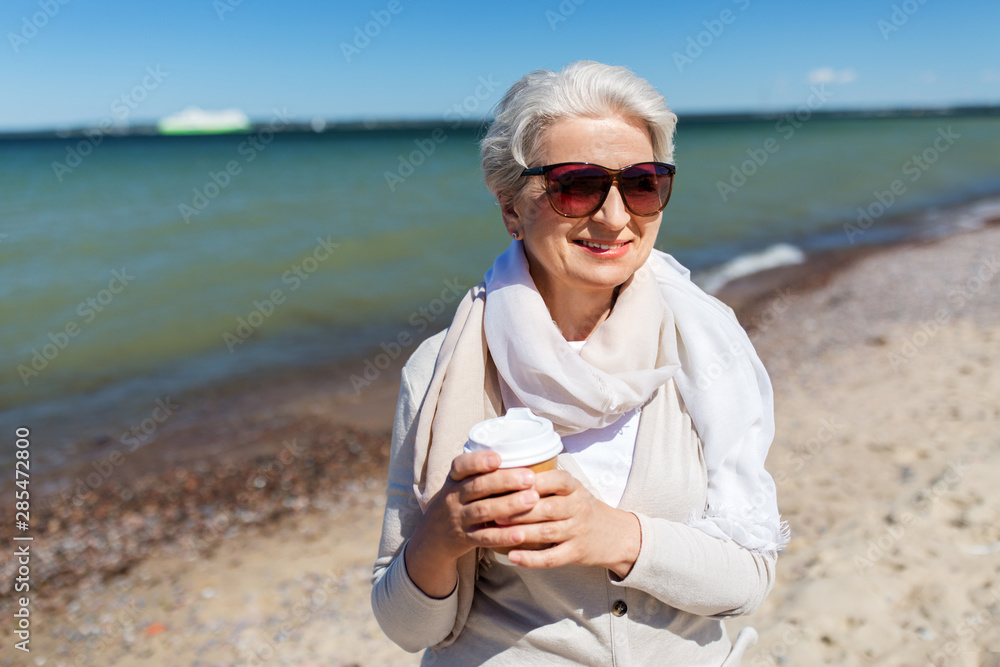 people and leisure concept - senior woman in sunglasses drinking takeaway coffee on beach in estonia