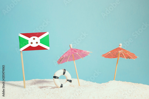 Miniature flag of Burundi on beach with colorful umbrellas and life preserver. Travel concept, summer theme. © sezerozger