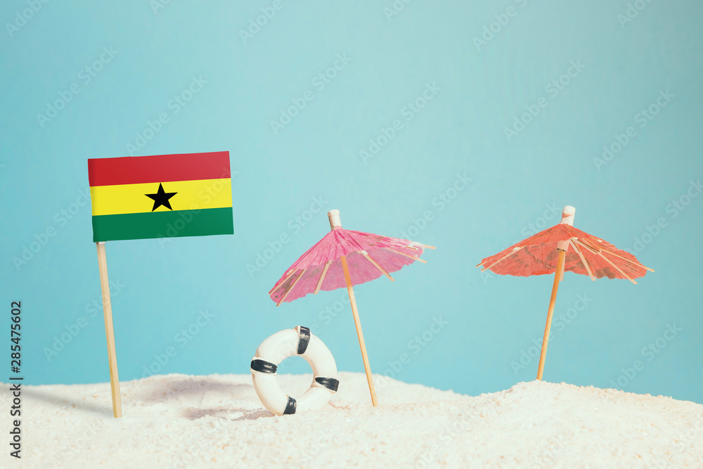 Miniature flag of Ghana on beach with colorful umbrellas and life preserver. Travel concept, summer theme.