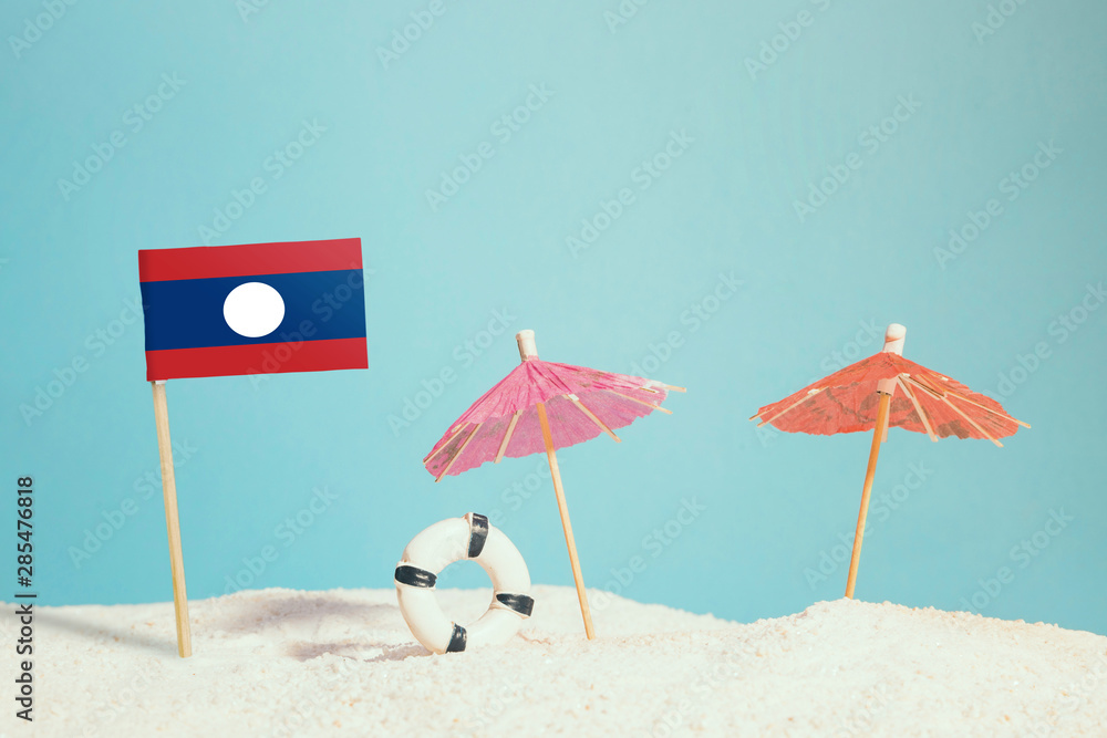 Miniature flag of Laos on beach with colorful umbrellas and life preserver. Travel concept, summer theme.