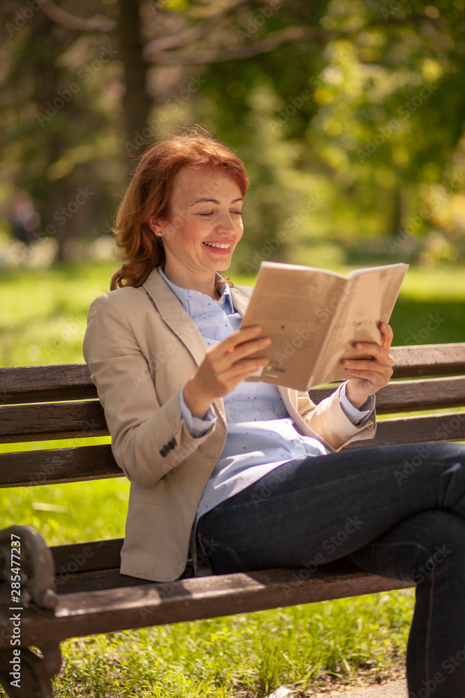 one mature interested woman, reading a book in a public while, sitting in bench. one mature interested woman, reading a book in a public while, sitting in bench.
