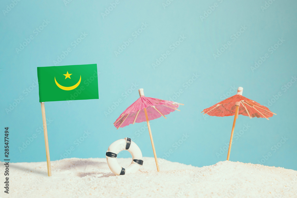 Miniature flag of Mauritania on beach with colorful umbrellas and life preserver. Travel concept, summer theme.