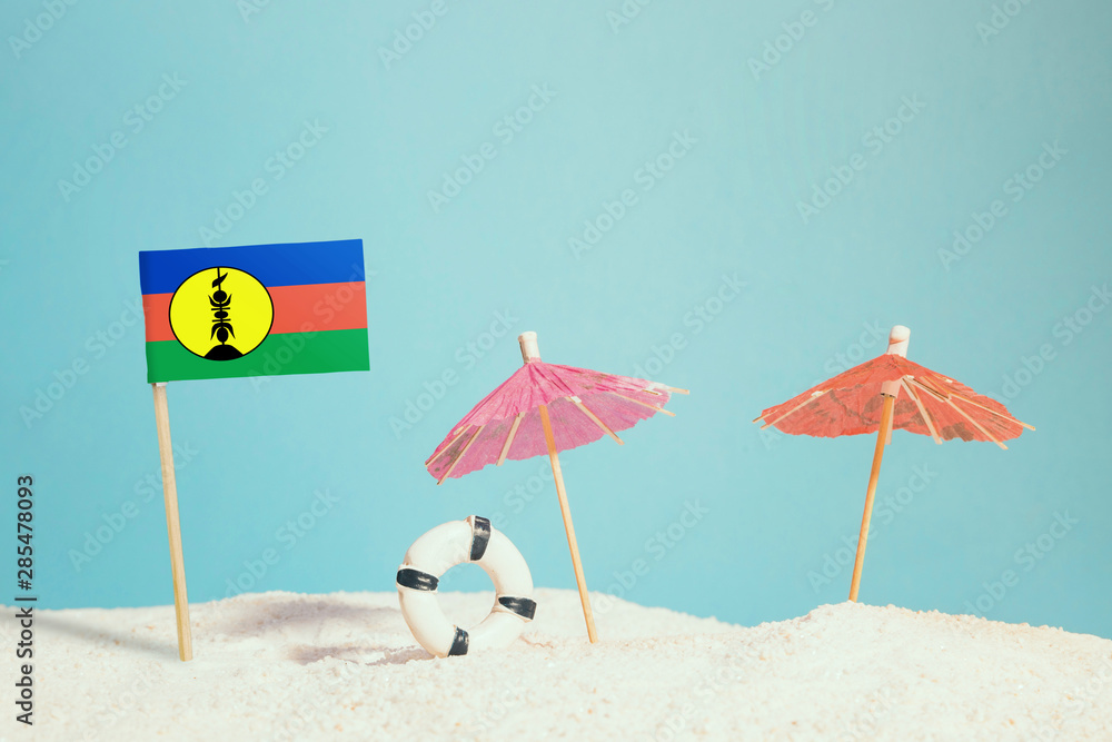 Miniature flag of New Caledonia on beach with colorful umbrellas and life preserver. Travel concept, summer theme.