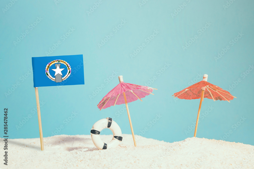 Miniature flag of Northern Mariana Islands on beach with colorful umbrellas and life preserver. Travel concept, summer theme.