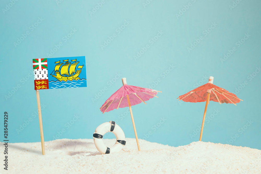 Miniature flag of Saint Pierre And Miquelon on beach with colorful umbrellas and life preserver. Travel concept, summer theme.