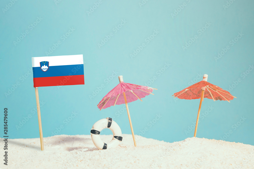 Miniature flag of Slovenia on beach with colorful umbrellas and life preserver. Travel concept, summer theme.