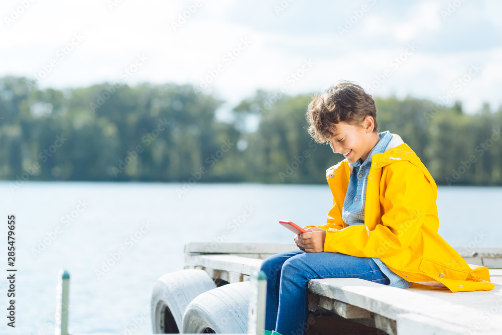 Smiling boy reading message on phone sitting near river