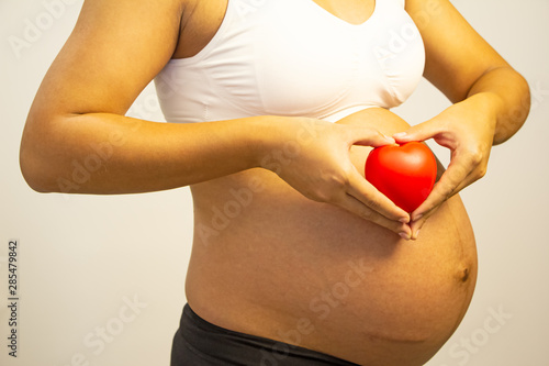 The pregnant woman holding small red heart in front of her striped belly,love and care for new born