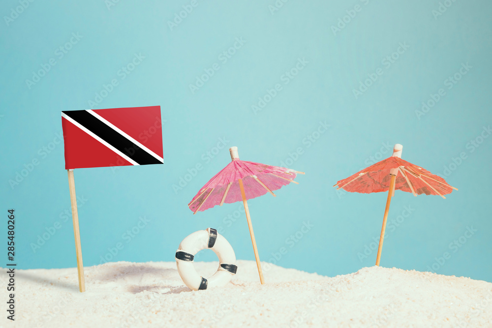 Miniature flag of Trinidad And Tobago on beach with colorful umbrellas and life preserver. Travel concept, summer theme. Miniature flag of Trinidad And Tobago on beach with colorful umbrellas and life preserver. Travel concept, summer theme.