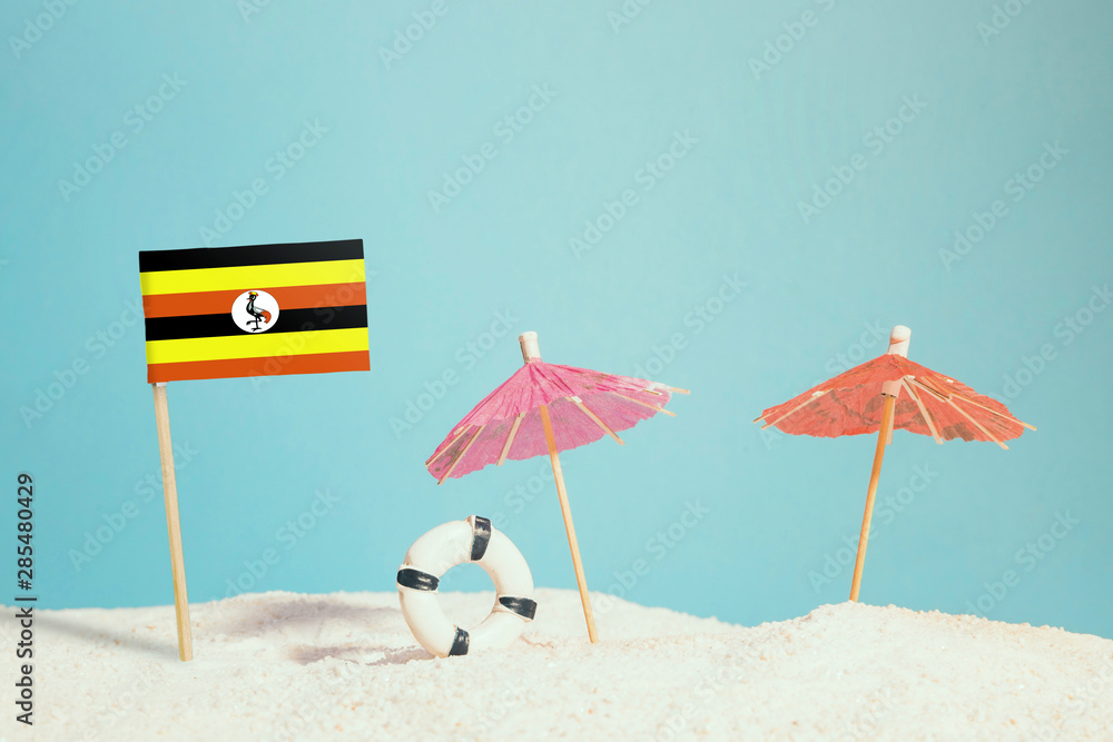 Miniature flag of Uganda on beach with colorful umbrellas and life preserver. Travel concept, summer theme.