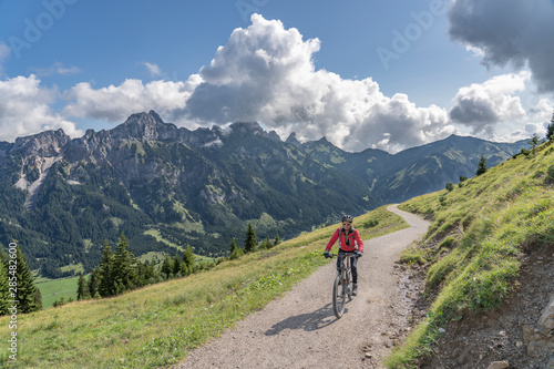 nice and active senior woman, riding her e-mountain bike in the Tannheim valley , Tirol, Austria, with the village of Tannheim and famous summits Gimpel and Rote Flueh