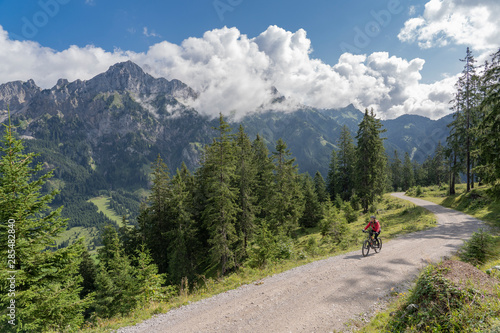 nice and active senior woman, riding her e-mountain bike in the Tannheim valley , Tirol, Austria, with the village of Tannheim and famous summits Gimpel and Rote Flueh