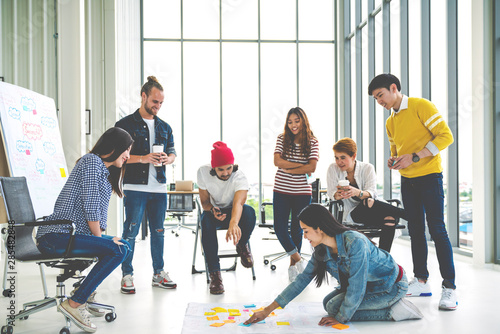 Young creative diverse group meeting and looking at project plan lay out on floor discuss or brainstorm business strategy with post note. Workshop for startup team in modern office. Happy workplace.