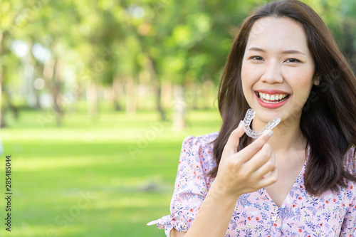 close up young beautiful asian woman smiling with hand holding dental aligner retainer (invisible) at outdoor nature park and garden background for beautiful teeth and dental treatment course concept