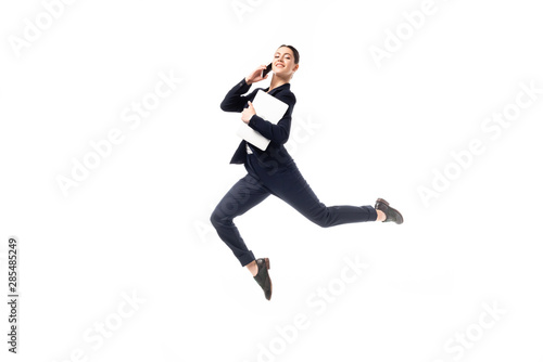 positive businesswoman talking on smartphone and holding laptop while levitating and looking at camera isolated on white