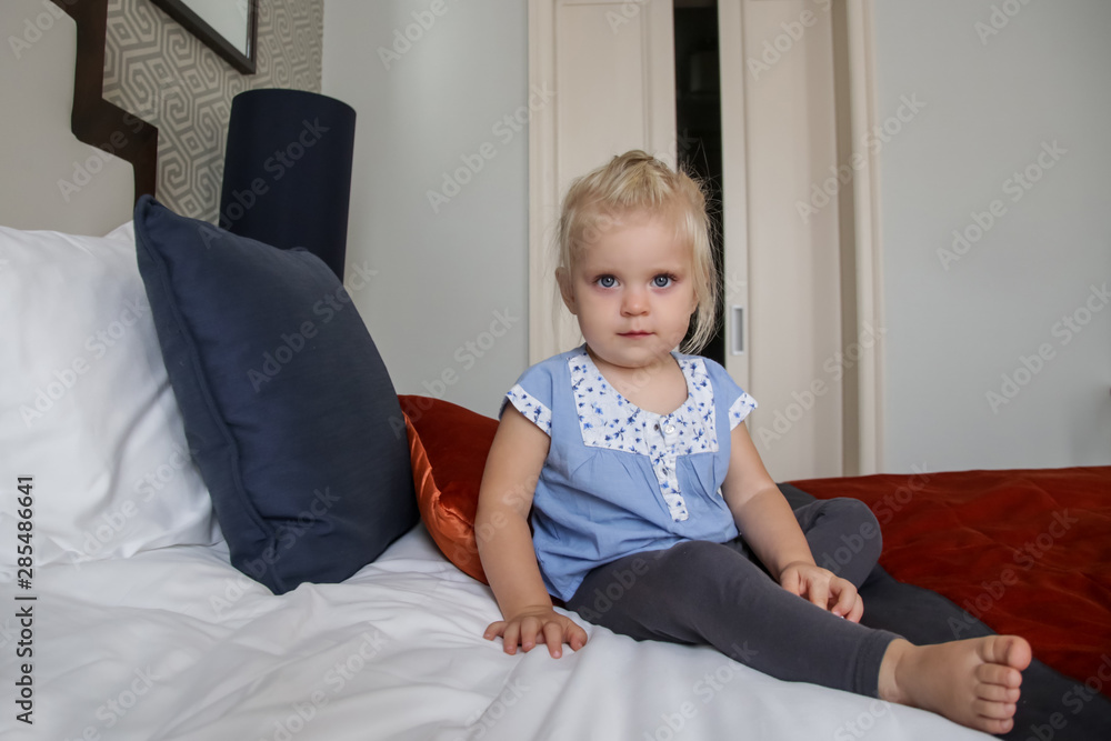 Fototapeta premium Cute blonde toddler girl in blue cotton shirt sitting on the bed 