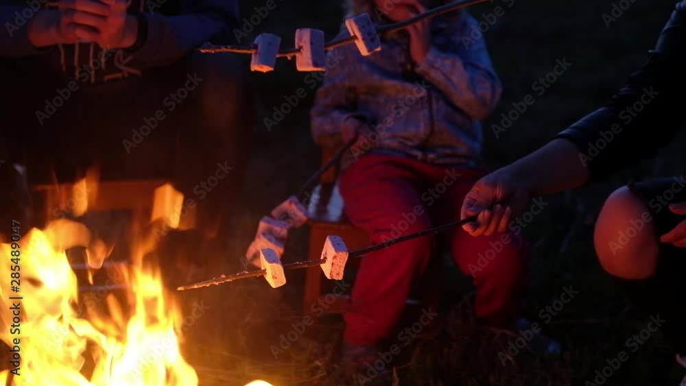 The European family sits around a blazing hot fire and fry square sweet marshmallows on long thin wooden twigs. Evening family gatherings by the flame in nature close-up. The European family sits around a blazing hot fire and fry square sweet marshmallows on long thin wooden twigs. Evening family gatherings by the flame in nature close-up.